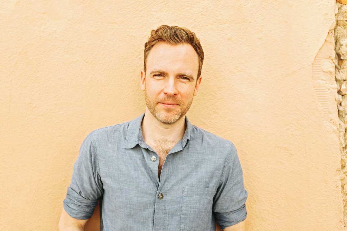 Portrait of Darren Wall, standing in front of a pale ochre wall.